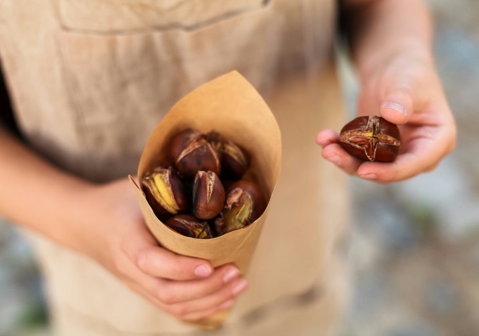 Person holding a brown paper cone filled with roasted chestnuts, with one chestnut held in the other hand.