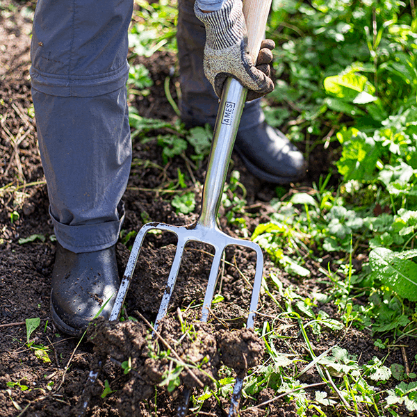 A person wearing gloves and boots uses a Digging Fork - Stainless Steel, crafted from FSC® certified ash wood, which comes with a 15-year guarantee, to dig soil in a green patch.