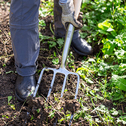 A person wearing gloves and boots uses a Digging Fork - Stainless Steel, crafted from FSC® certified ash wood, which comes with a 15-year guarantee, to dig soil in a green patch.