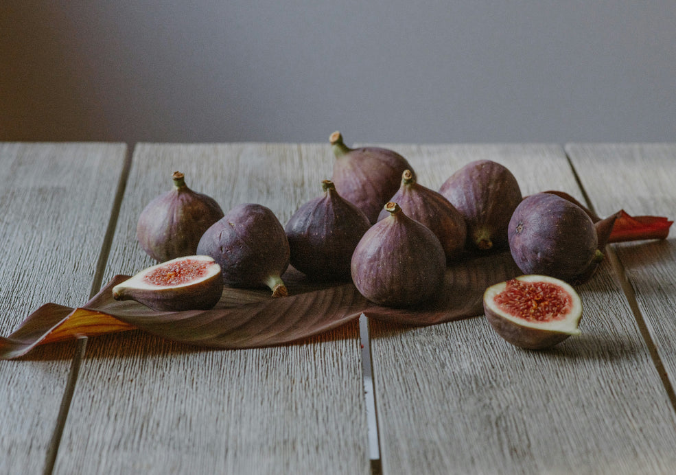 A group of fresh figs, including whole and halved, are arranged on a large leaf on a rustic wooden table.