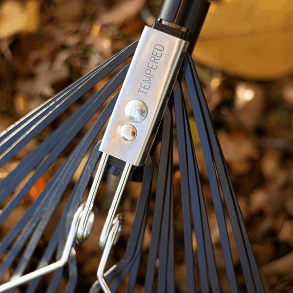 Close-up of a Flat Tine Leaf Rake made from Carbon Steel with a TEMPERED label, featuring an FSC certified ash wood handle, positioned over autumn leaves, ideal for clearing garden debris.