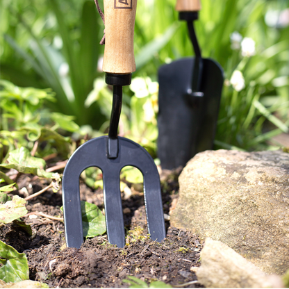 A Hand Fork - Carbon Steel, featuring an elegant ash wood handle, is positioned in soil among green plants and rocks alongside a matching trowel.