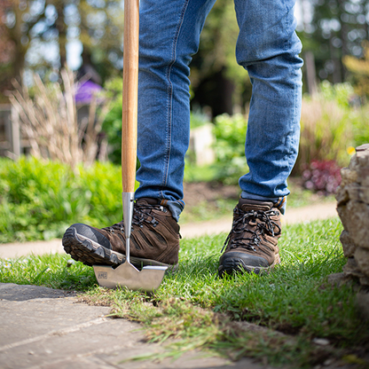 A person in jeans and boots skilfully maneuvers a Lawn Edger - Stainless Steel, expertly defining the edges of a lush lawn.