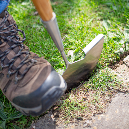 Wearing brown boots, an individual deftly handles the Lawn Edger - Stainless Steel with an ash wood handle, carefully trimming the grass along the path.