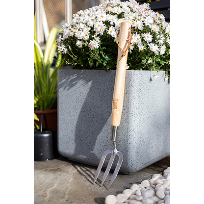 A Midi Hand Trowel with an FSC ash wood handle leans against a large grey planter filled with blooming white flowers. Pebbles encircle the base on a patio, enhancing the serene garden scene.