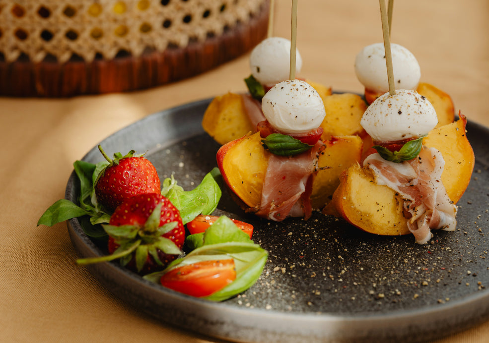 A plate with skewered mozzarella, peach, basil, and prosciutto slices, next to fresh strawberries and cherry tomatoes.