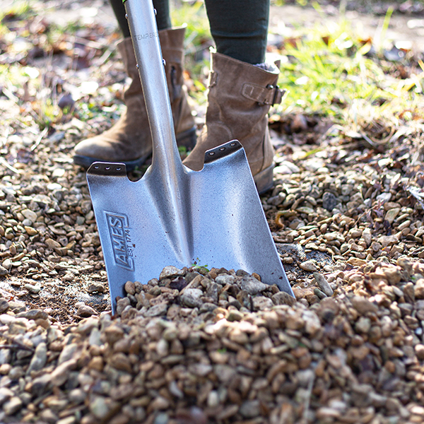 A person wearing boots uses the Square Mouth Shovel - Carbon Steel, featuring an ergonomic handle, to move gravel efficiently.