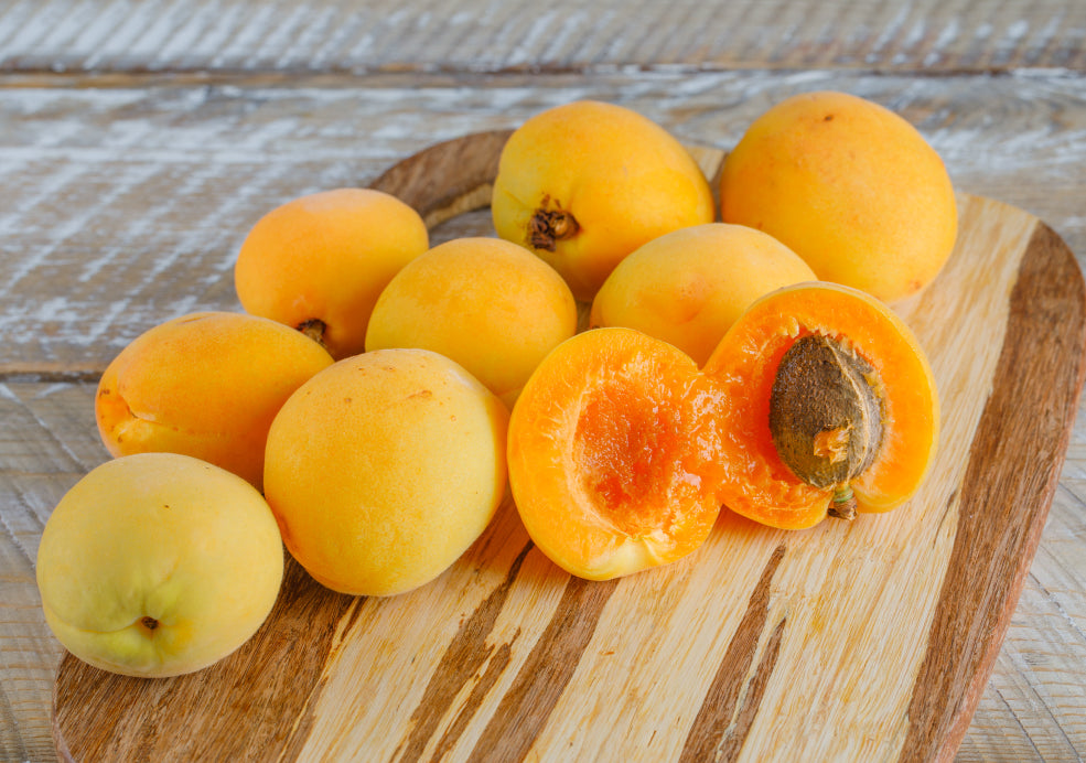 A group of whole apricots and one halved apricot with a visible pit on a wooden cutting board.