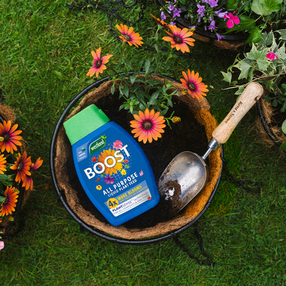 A container of Westland Boost All Purpose Liquid Plant food 1L and a gardening trowel are placed in a soil-filled planter, enhanced by PlantSense Technology, surrounded by flowering plants on the grass.