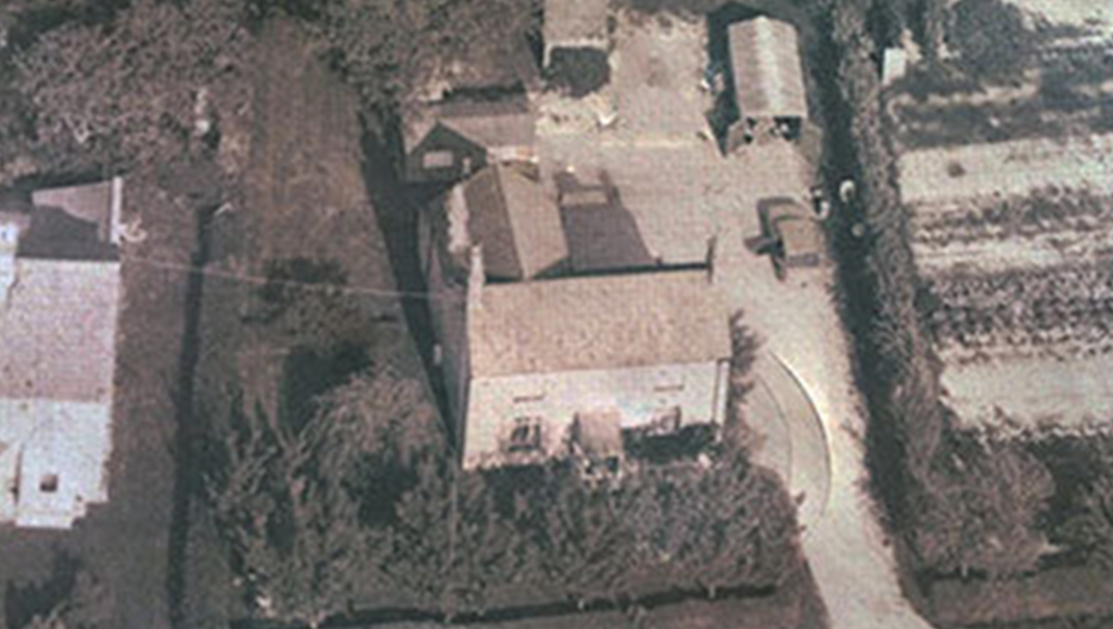 Aerial view of a rural property with a house, outbuildings, driveway, and surrounding vegetation.