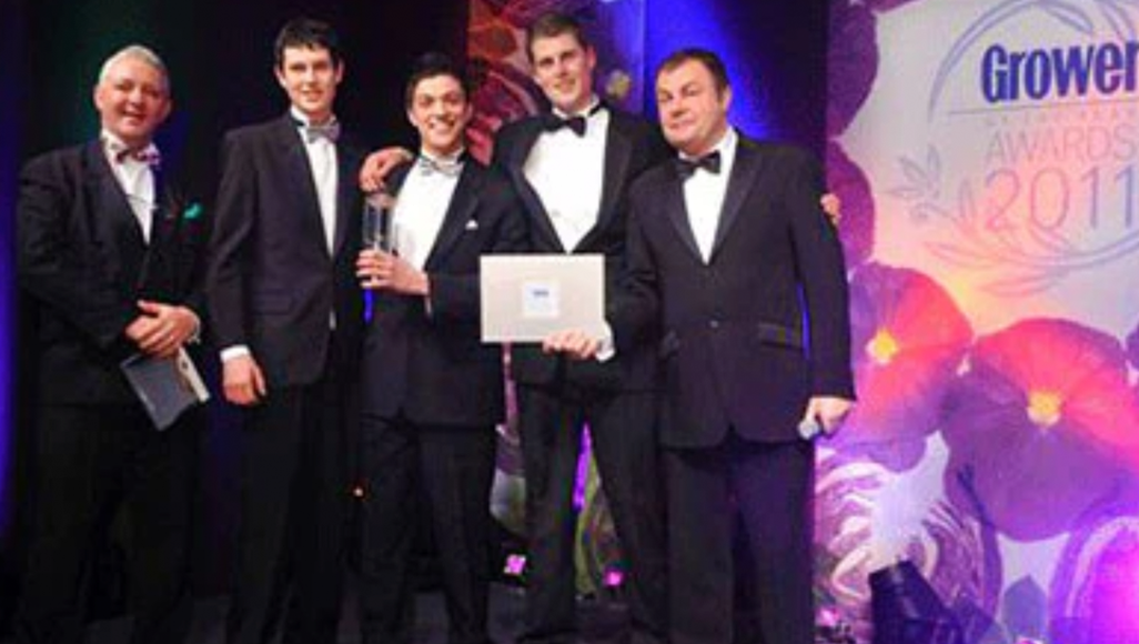 Five men in tuxedos stand on stage at an award ceremony, holding a trophy and certificate, with a backdrop displaying 2011 and floral graphics.